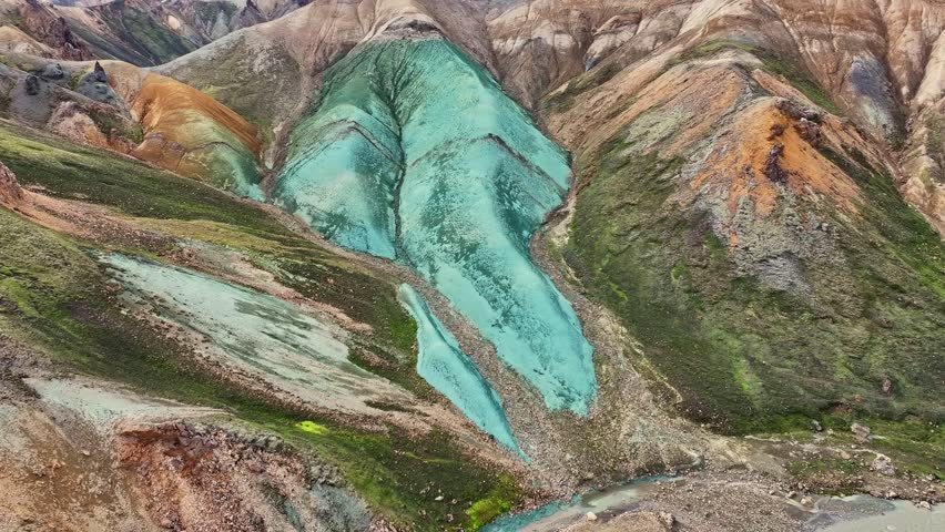Aerial drone fast backward movement at medium altitude, over Grænihryggur, the green rock, the river, and the valley in Landmannalaugar, Iceland, highlighting medium tones of orange and green.
