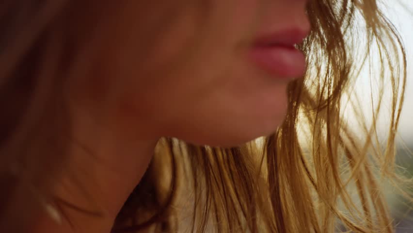 CLOSE UP, LENS FLARE: Gentle summer wind blowing through woman's long wavy hair. Rear view of beautiful young brunette, illuminated with amazing golden light, is walking towards the setting summer sun