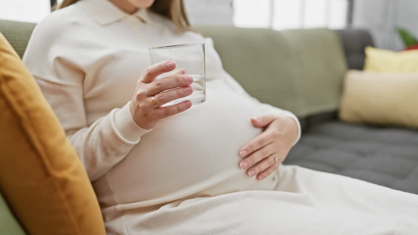 A pregnant hispanic woman holding a glass sits on a couch indoors, depicting maternity and anticipation. Royalty-Free Stock Footage #3432555453