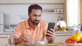 Indian middle aged man busy using mobile phone while eating lunch on dining table at home - concept of modern lifestyles, internet distraction and social media sharing