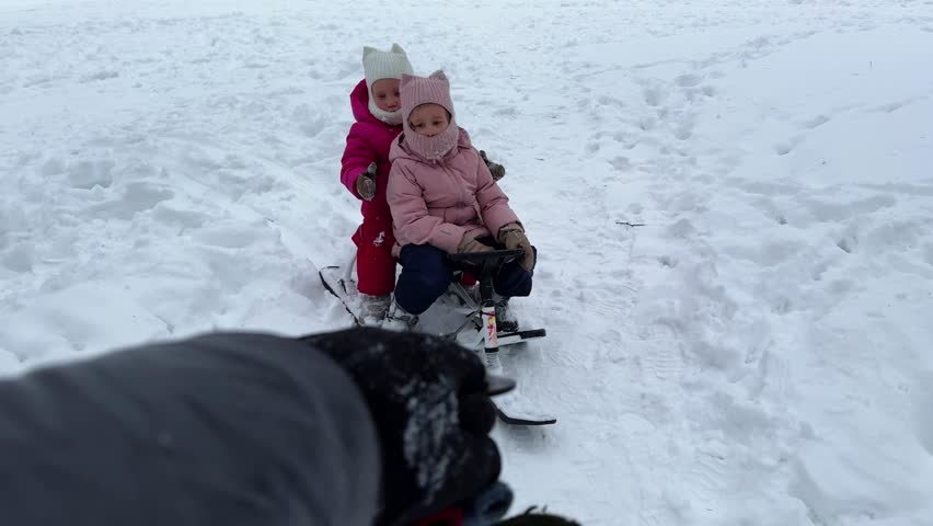 Dad is carrying two little twin daughters by a string on a snow scooter in the snow, first-person 