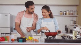 Indian couple at home preparing food by watching recipe on mobile phone app at kitchen during weekend - concept of relationship bonding, technology and teamwork