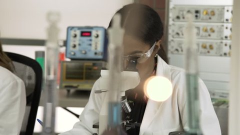 Scene tracks slowly past laboratory glassware and lights in the foreground to a young African American female scientist or medical student using a microscope and making notes about what she sees.