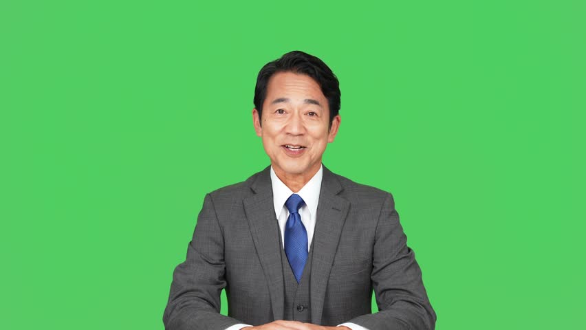 An middle-aged Asian man in a suit talking to the camera. green background for chroma key composition. announcer. news caster.