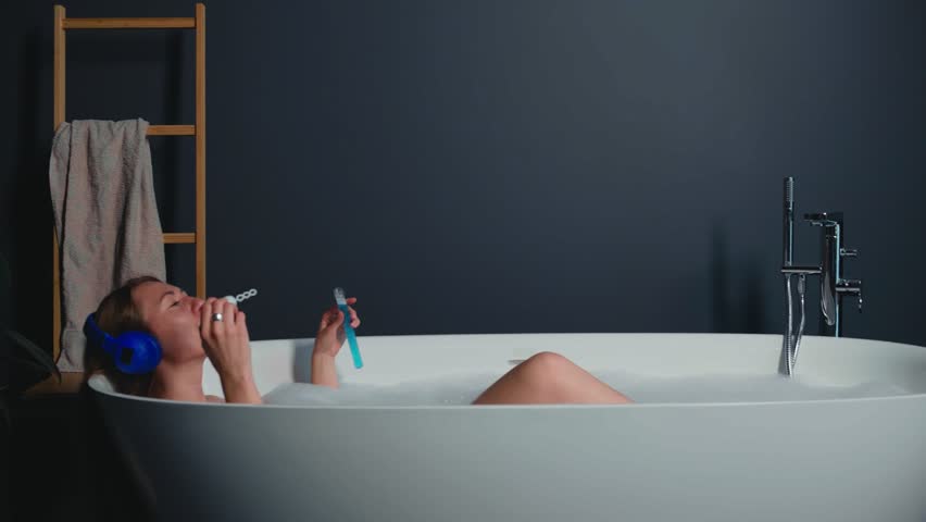 Happy serene relaxed beautiful young woman in headphones lying in modern stylish bath having fun blowing soap bubbles.