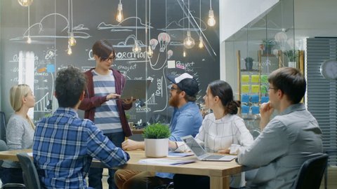 Charismatic Asian Team Leader Shows Laptop to a Diverse Group of Talented Young Developers, They Start Discussion at the Meeting Table. Creative People in Stylish Office Environment. 4K UHD.