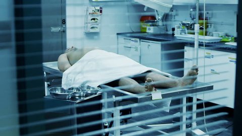The lifeless naked corpse of a young mixed race male is laid out on the autopsy table, ready for the medical examiner to begin his work. He checks the toe tag and starts to make notes.