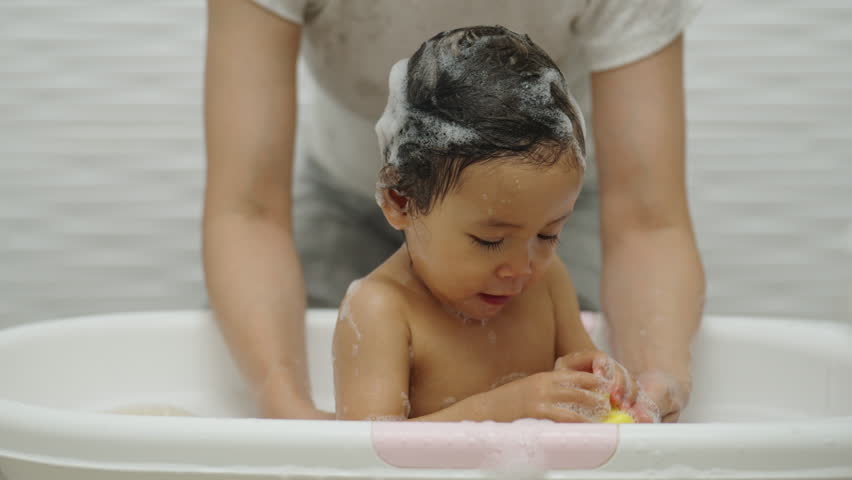 mother bathing her cheerful infant baby with foam bubbles and playing rubber duck in a bathtub