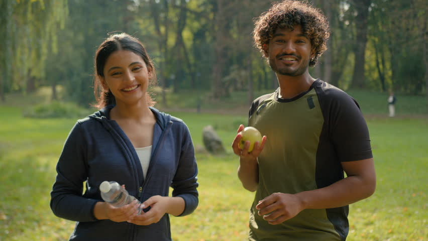 Indian man and woman relaxing after sports together in park outdoors Arabian fitness couple friends in city sport happy people showing holding apple fresh healthy fruit and bottle of water lifestyle Royalty-Free Stock Footage #3432881409