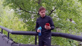 A male runner takes a break to drink water while using an app on the smartphone.