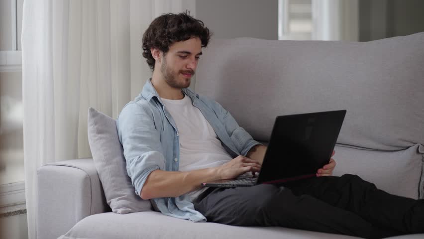 Relaxed serious millennial caucasian guy student freelancer using laptop device leaning on sofa at home office, focused entrepreneur working distantly typing on notebook in apartment
