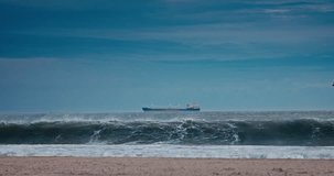 Seascape of rolling sea waves on the beach coast and sailing cargo ship in the water toward industrial harbour