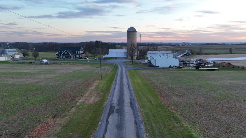 Dawn over a rural farm lane leading to farmhouses and a silo among open fields in American countryside. Aerial during sunrise.