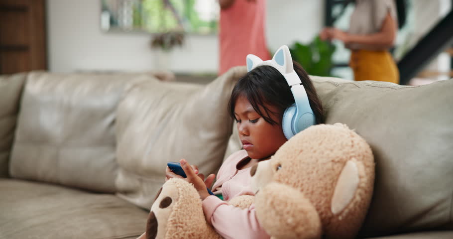 Child, phone and headphones with parents in argument, disagreement or divorce on living room sofa at home. Little girl or daughter playing on mobile smartphone with teddy bear in family conflict