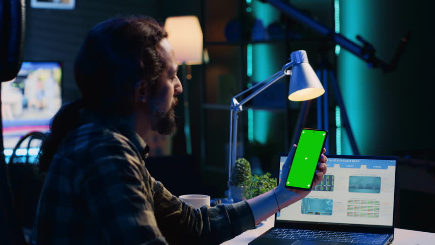 Teleworker holding green screen smartphone, analyzing statistical charts on laptop in living room. Self employed man checking business revenue numbers on mockup mobile phone
