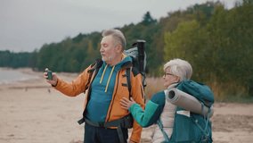 Senior couple tourists bloggers taking photo video on smartphone standing on sandy riverbank. Hikers old gray haired man and woman family travelling carrying backpacks with touristic equipment.
