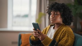Young woman sitting with phone. African American woman hold in hand use mobile phone typing messages. Girl relax spend free spare time laughs in room. Communication on social networks, rest concept. - Powered by Shutterstock - Get 15% off with code: PIKWIZARD15