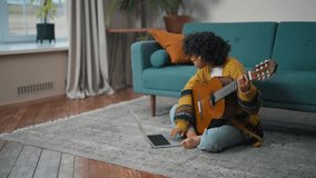 Young girl learning to play guitar. African american teenager girl study to playing the guitar see in laptop sitting on floor at living room. Online-education, musician art, hobby practice concept.