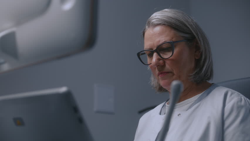 Mature female doctor looks at monitor of ultrasound machine while doing sonography diagnostics. Medical specialist performs check up or examination in modern clinic using advanced equipment. Portrait. Royalty-Free Stock Footage #3433472909