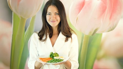 Pretty girl in a white robe holds a large plate of boiled vegetables and pushes it forward. Brunette winks. Healthy and proper nutrition. Vegetarian food.