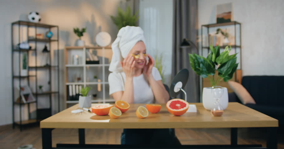 Happy woman in white towel on her head which applying golden collagen eye patches and looking posing on camera with sincerely smile