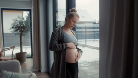 Anxious pregnant woman standing next to the window and touching her abdomen