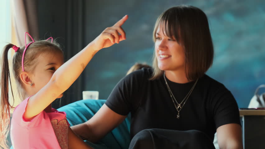 Young mother and small daughter spend time together. Girl points somewhere with finger telling story to woman sitting in cafe