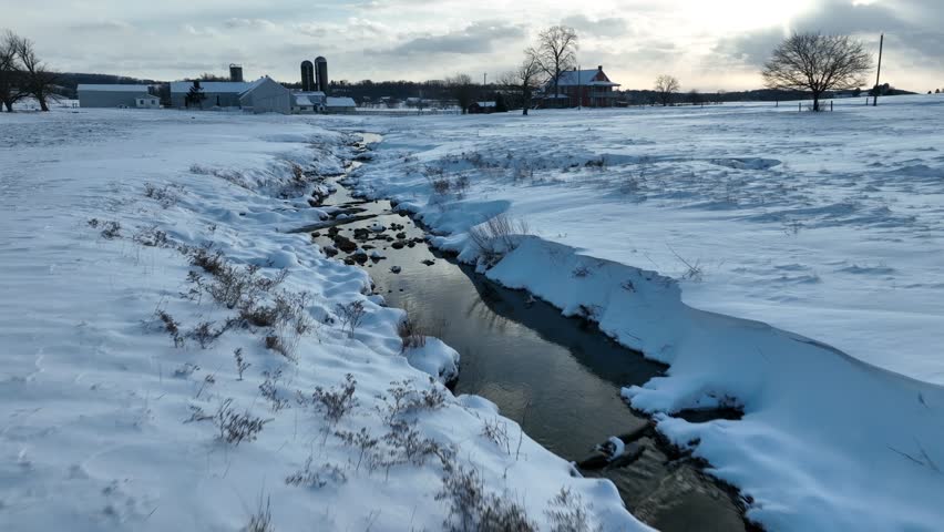 Snowy landscape with a meandering stream near a farm under a cloudy sky. Low aerial glide in rural USA with snow covered farmland.