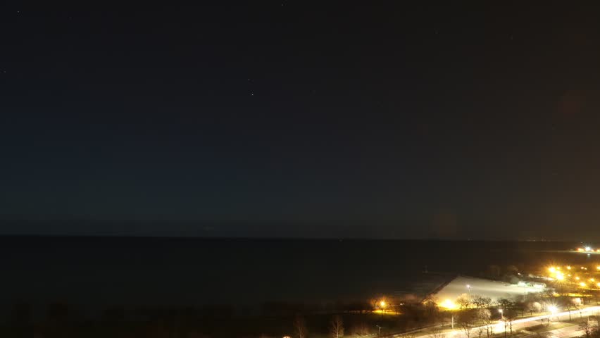 Night time-lapse of star trails over Lake Michigan in Chicago with airplanes and clouds leaving their marks in the black night sky above and water below.