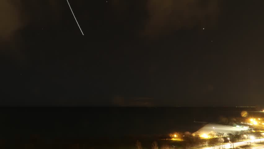 Night time-lapse of star trails over Lake Michigan in Chicago with airplanes and clouds leaving their marks in the black night sky above and water below.