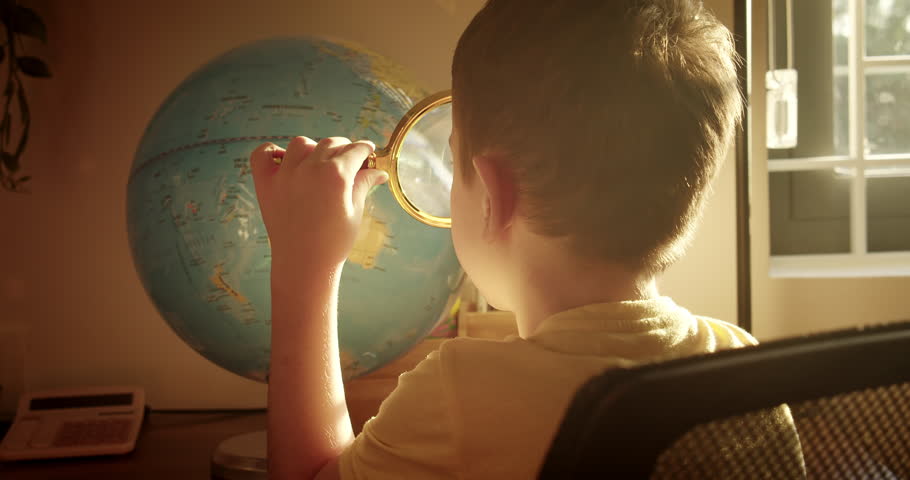 Kid in morning light sunset, cute little boy looking at globe through a magnifying glass, technology for schoolchildren. View from back Child is studying while sitting at home in rays sunset in room.