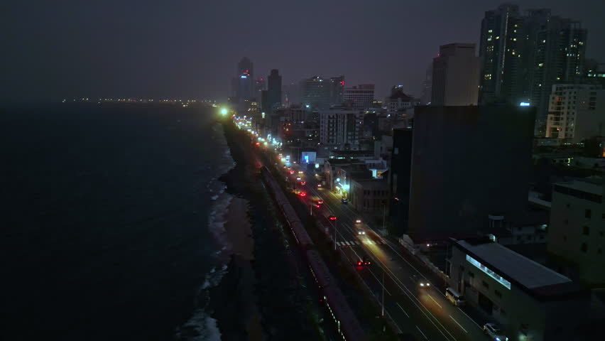 Aerial night view, Colombo skyline illuminated, vehicles move on coastal road, waves crash against shore, urban Sri Lanka lit up, cityscape after dark with glowing lights, seaside metropolis vista.
