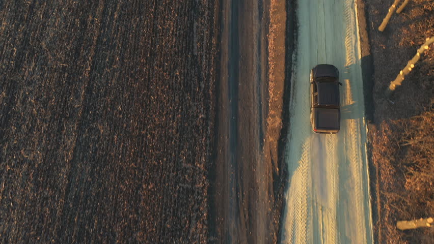 Flying over off road vehicle moving on route near farmland. Aerial shot of pickup truck driving through rural path near ploughed field. Black car moving along road near plowed meadow at autumn