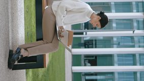 Business woman starting online meeting putting on earbuds outdoor. Smart confident girl conducting web conference on laptop sitting bench near office vertical view. Manager using notebook for work.