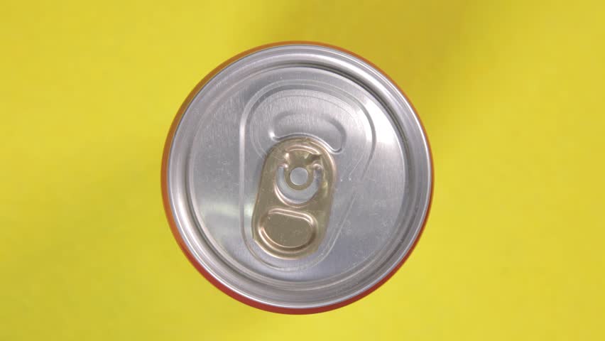 Above close-up shot: a hand opening a tin can, unleashing a burst of effervescent gas from a rich dark liquid carbonated drink, then taking the container away.