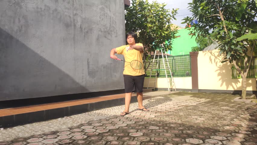 A good person wearing a yellow shirt and black track pants takes advantage of their free time by engaging in light exercise. During the hot afternoon, they play badminton in the yard next to the house