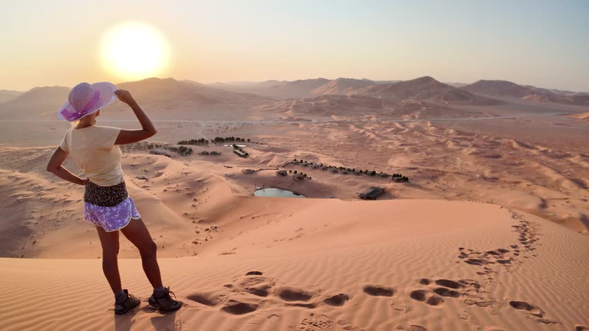 A woman at sunset on the sands of Oman's Empty Quarter at sunrise, relishing the dawn spectacle amidst golden dunes, a vivid contrast of colors in the tranquil desert morning. Royalty-Free Stock Footage #3434438171