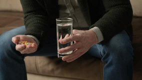 Hands with pills, close-up. Senior man hands holding medical pill and glass of water. Man taking medication cure pills, sitting on couch. Medicine healthcare therapy concept, 4k slow motion