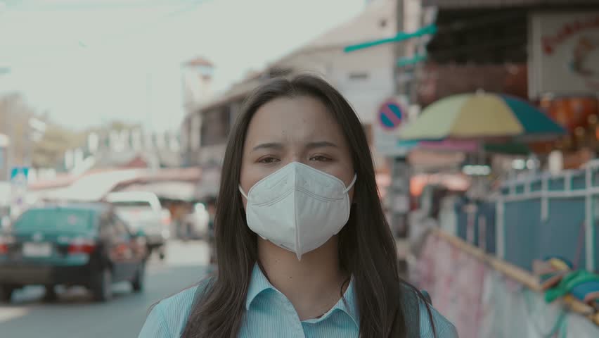 A sick Asian woman wears a mask to protect against PM 2.5 dust coming from a busy city and air pollution. Smog, dust and bad weather. Pollution. Covid. Bad air.
