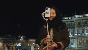 A beautiful Asian girl takes a video on her phone on a three-axis stabilizer on the night streets of a winter city. A young blogger sets up her mobile phone for an online broadcast from city square
