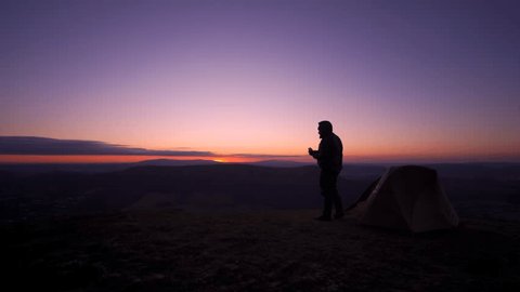 An explorer eating breakfast at dawn on a hill summit beside a wild camping tent in gorgeous morning light
 Video stock