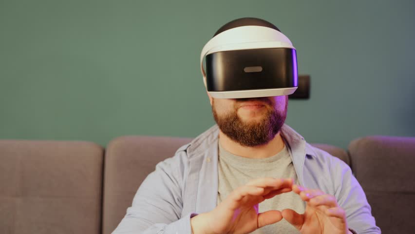 Close-up portrait of a Caucasian young man wearing VR glasses, moving his hands in the air and making some gestures like scaling and swiping photos in virtual reality in his living room at home