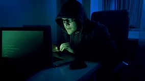 Funny video of young male pretends to be hacker. Geeky man in hood working on laptop at night