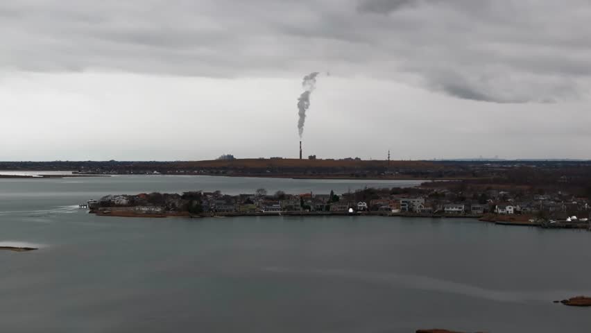 An aerial view over a marsh on a cloudy day with gray skies by Freeport, NY. The camera dolly in towards a power plant with a smokestack blowing out white steam into the atmosphere in the distance.