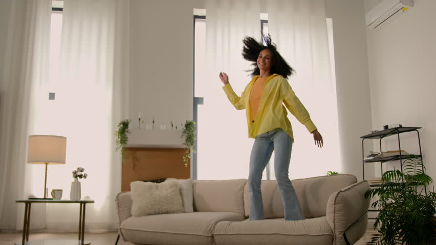 Active funny crazy African American girl young happy woman jumping on sofa couch in living room excited female jump dance loud music home party rejoicing carefree joyful lady weekend vacation freedom Royalty-Free Stock Footage #3434739393