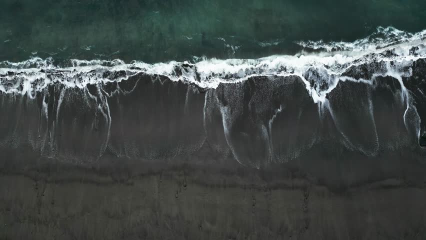 A wave hitting the black sand beach, showcasing the contrast of white foam against the dark shoreline, aerial view