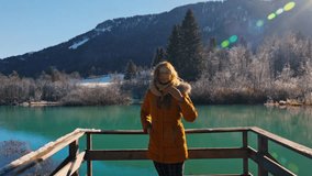 4K video of a happy young caucasian female standing near the lake in the natural reserve - Zelenci, Slovenia. Looking in the camera, smilling and observing scenery with yellow winter jacket.