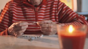4K video of an elderly women holding the holy rosary and praying next to the Holy Bible. Indoor scene with candle light.
