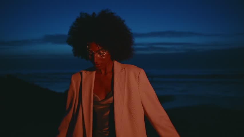 Fashion model dancing raise hands at dark seashore. Closeup seductive african american posing spin on night beach. Festive lady enjoy summer look camera in pink light. Party woman with glitters makeup