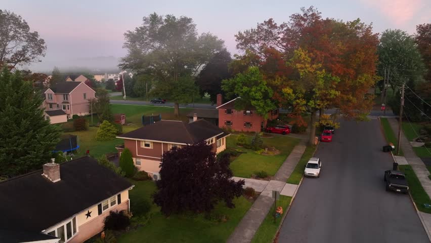 Foggy neighborhood during autumn sunrise. Aerial rising shot revealing colorful trees and large American homes.